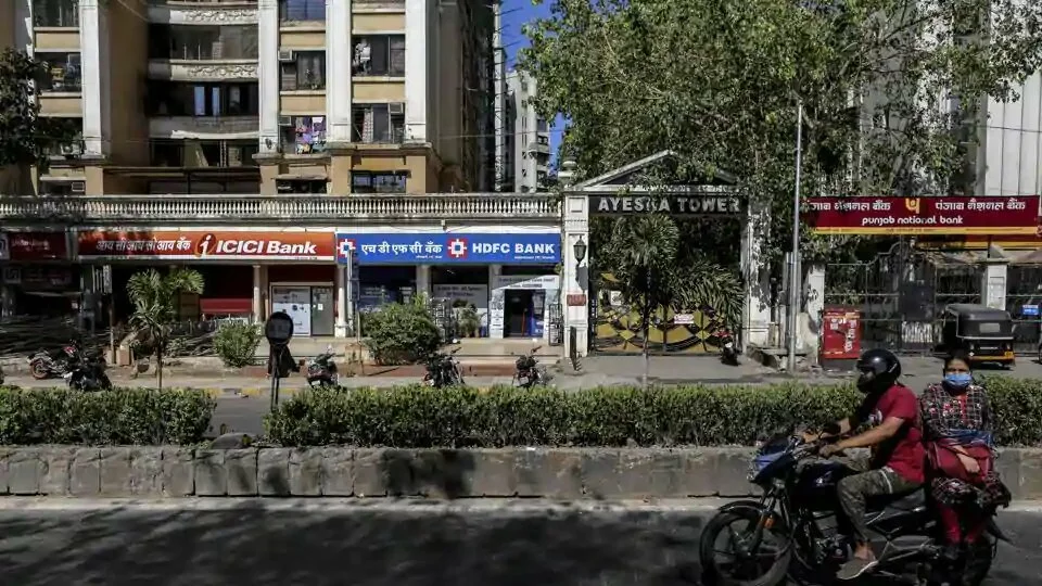 A motorcyclist and passenger travel past branches of ICICI Bank Ltd., HDFC Bank Ltd. and Punjab National Bank (PNB) on a near-empty street in Mumbai.