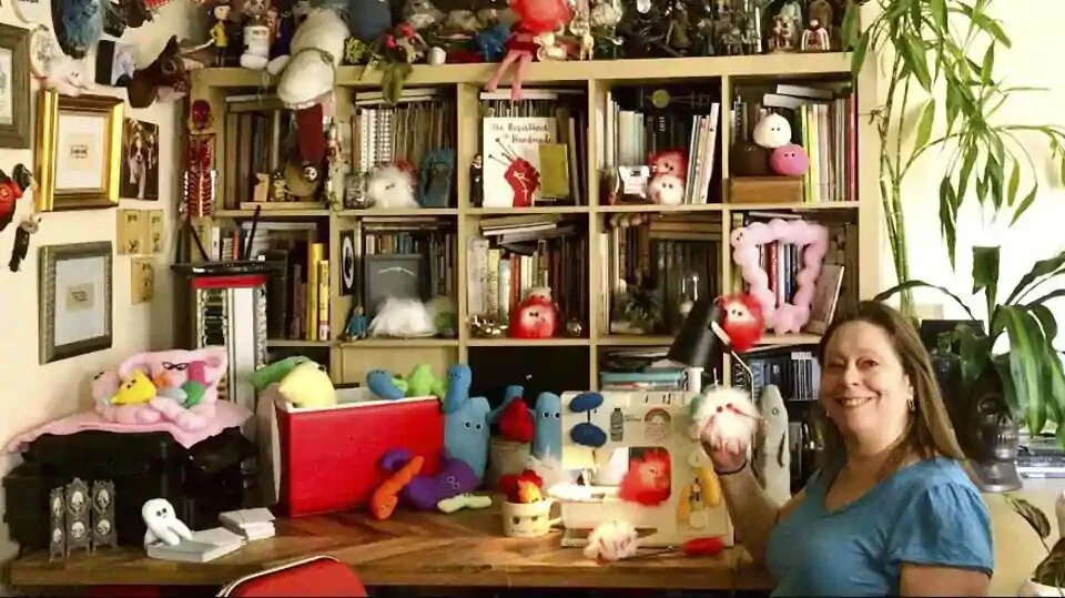 This July 25, 2020 photo shows Amy Micallef posing with her craft creations at her home. While Broadway stages remain dark due to the pandemic, Broadway workers are concentrating on side hustles. Micallef, a Broadway seamstress who has worked in the wardrobe departments of “Hamilton,” “Waitress” and “Frozen,” makes gleeful representations of COVID-19 for sale, complete with a pair of eyes and faux fur.